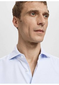 Svetlo modra srajca z ovratnikom na konico, z zapenjanjem na gumbe in gladko teksturo. Ima en žep, krojena fit oblika in brez vidnih vzorcev.