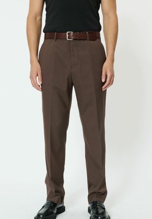 Muž oblečený v hnedých mierne priliehavých nohaviciach, čiernom opasku, čiernej vtiavanej košeli a čiernych kožených mokasínach stojaci pred jednoduchým pozadím.