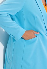 Blazer su misura in azzurro chiaro con una tasca singola e tre bottoni abbinati sul polsino, realizzato in un tessuto liscio.