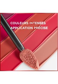 Applicateur de maquillage avec un embout doux et texturé, placé sur des échantillons de couleurs vibrantes et brillantes dans des teintes de rose, rouge et nude sur une surface lisse.