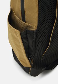 Zaino con esterno in tela beige, dettagli in rete, due scomparti laterali e una tasca nera con zip per riporre gli oggetti. Dettaglio in tessuto testurizzato evidente.