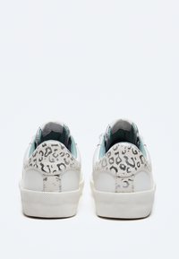 Zapatillas blancas con acentos de estampado de leopardo en el talón, suela de goma texturizada y un forro interior azul claro. Diseñadas para uso casual.