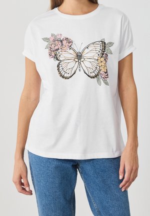 Mujer con camiseta blanca con un gran diseño de mariposa y flores rosas, combinada con jeans azules, de pie contra un fondo liso.