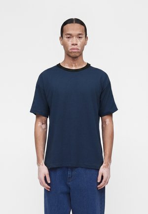 Άνδρας με λεύκη που φοράει μπλε σκούρο T-shirt και μπλε τζιν, στέκεται μπροστά σε απλό λευκό φόντο, κοιτάζοντας ευθεία μπροστά.