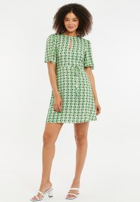 Vihreä kuvioitu mekko, jossa on lyhyet hihat, avonainen kaula-aukko ja vyötäröllä solmittava vyö. Kuvio koostuu vuorottelevista muodoista mustassa ja valkoisessa.