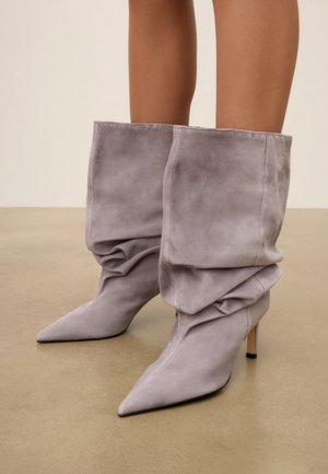 Femme portant des bottes à talons hauts en daim gris, à bout pointu et effet décontracté, sur un sol beige.