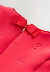 Punast kangast riietus, millel on tekstureeritud pind, milles on reljeefsed tähed ja kuldne lukk. Kaunistatud vastava punase paelavõruga.