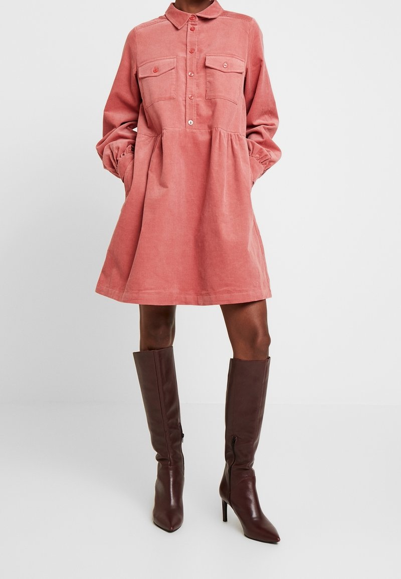 Robe-chemise en velours côtelé rose corail avec poches poitrine et manches longues, portée avec des bottes en cuir marron foncé montant jusqu'au genou et à talons.