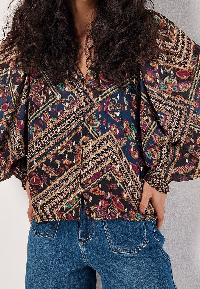 Chemisier boutonné en tissu foncé avec des motifs floraux et géométriques colorés, doté de manches larges et de poignets élastiques. Associé à un jean en denim.