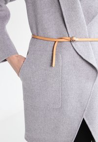 Persoon met een lichtgrijze wollen jas met grote voorzakken en een dunne tan leren riem die losjes om de taille is geknoopt.