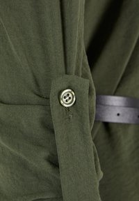 Gros plan sur un tissu vert olive avec un détail de sangle boutonnée et une vue partielle d'une ceinture noire en arrière-plan.