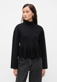 Svart turtleneck-genser med flagrende nederst og lange ermer, laget av strukturert strikket stoff. Kombinert med høyh teide mørke jeans.