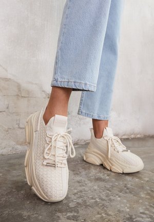 Feet wearing chunky off-white knit sneakers with thick soles and cream laces, paired with light blue cropped denim jeans on concrete floor.