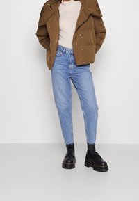 Chaqueta de plumas marrón, suéter ligero, jeans azul de cintura alta y botas de tobillo negras con cordones y suela gruesa. Atuendo urbano simple y moderno.