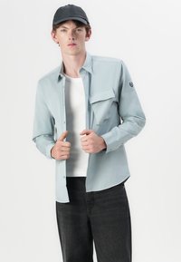 Camisa azul clara de botões com um bolso no peito, usada sobre uma t-shirt branca. Jeans escuros e um boné preto completam o visual.