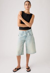 Γυναίκα όρθια με τα χέρια σταυρωμένα, φορώντας μαύρο αμάνικο τοπ, ανοιχτό μπλε τζιν σορτς μέχρι το γόνατο και μαύρα σαγιονάρες, μπροστά σε απλό φόντο.