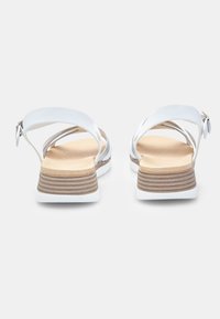 Valkoinen sandaali, jossa on ristikkäinen hihnasuunnittelu ja säädettävä solki. Varustettu pehmeällä ruskealla pohjalla ja pinottu beige korko. Tarjoaa tyylikkään, minimalistisen ilmeen.