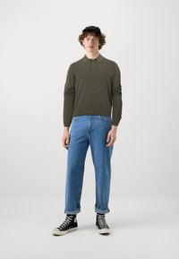 Camisa polo de manga larga verde oliva, vaqueros de corte relajado azules y zapatillas negras de caña alta. El fondo es blanco liso.