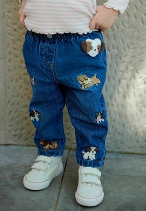 Kleinkind trägt blaue Jeans mit Hundemotiven, hellrosa gestreiftes Shirt und weiße Klett-Sneaker und steht auf gefliestem Boden.