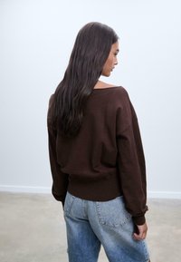 Brauner oversized Pullover mit weiter Ausschnitt und geripptem Saum, kombiniert mit hochgeschnittenen, hellblauen Jeansshorts in lässigem Schnitt.