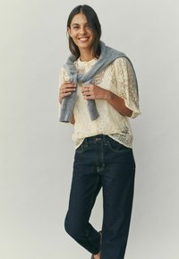 Mežģīņu blūze gaiši dzeltenā krāsā ar pelēkas krāsas šalli, kas sasieta mezglā un pārmetta pār pleciem; kopā ar tumši zilu taisnu džinsu biksēm.