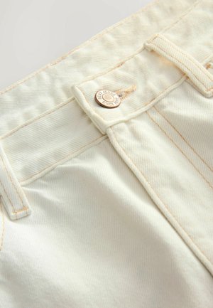 Weißer Denim-Stoff mit orangefarbigen Nähten, ausgestattet mit einem Metallknopf, auf dem "NEW JEANS SINCE 1982" eingraviert ist. Nahaufnahme der Designdetails des Taillenbunds.