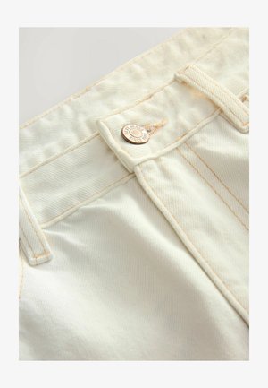 Baltas džinsu audums ar oranžiem diegiem, ar metāla pogu, uz kuras ir iegravēts "JAUNIE DŽINSI NO 1982. GADA". Tuvplāns uz jostas dizaina detaļām.