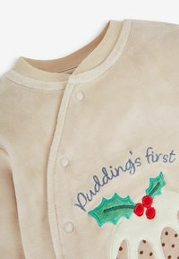 Beige színű baba bodyn, patentgombokkal és hímzett szöveggel: "Pudding első" egy karácsonyi puding felett, melyen fagyöngylevelek és piros bogyók találhatók.