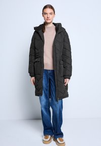 Giovane donna che indossa un lungo cappotto imbottito nero sopra un maglione beige, jeans blu e scarpe slip-on beige morbide, in piedi su uno sfondo neutro.