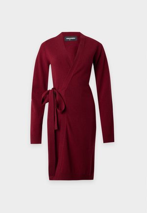 Vrouw met lang donker haar die een lange bordeauxrode cardigan, een grijs topje, een donkere spijkerbroek en bruine sandalen draagt, staand tegen een effen lichte achtergrond.
