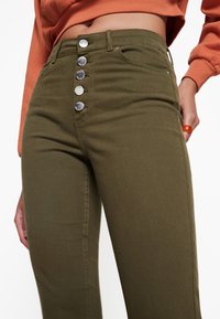 Jean taille haute vert olive avec une texture lisse, dotés d'une fermeture à cinq boutons à l'avant et de poches latérales. Les coutures sont visibles.