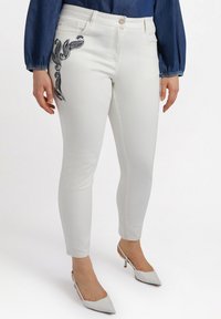 Jeans de mezclilla elástica blancos con un diseño ajustado, que presentan un acento de bordado de paisley negro en el muslo izquierdo. Combinados con zapatos de tacón blancos.