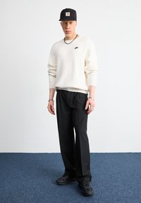 Valkoinen neulepaita mustalla Nike-logolla, mustat rentoa tyyliä olevat housut, mustat lenkkarit, lippalakki ja hopeiset asusteet. Sininen matto taustana.