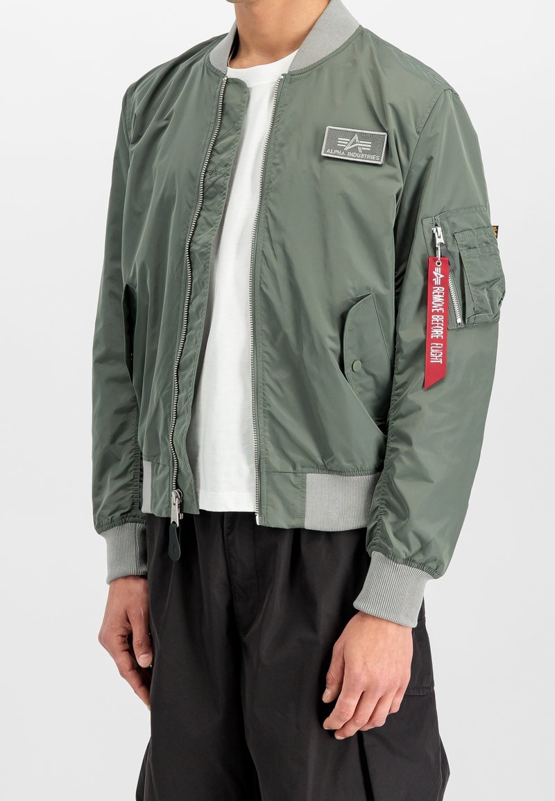 Personne portant une veste bomber vert olive non fermée avec des poignets côtelés gris, un t-shirt blanc, un pantalon noir et une étiquette rouge « Remove Before Flight » sur la poche de la manche.
