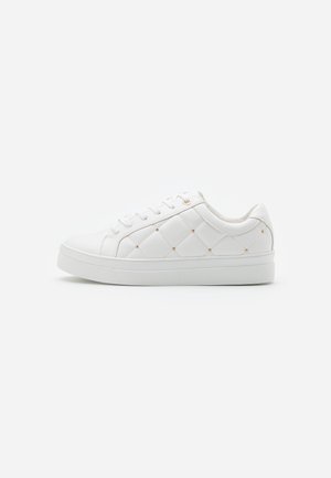 Witte lage sneaker met doorgestikte zijpanelen en kleine gouden studs, vetersluiting en platte rubberen zool op een effen achtergrond.