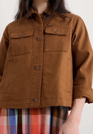 Kvinde iført en brun jakke med knapper og klaplommer over et flerfarvet ternet skørt, med ærmet rullet op på den ene arm.