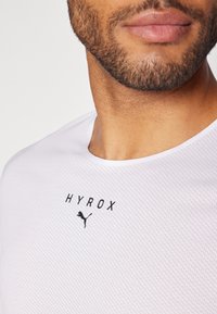 T-shirt de sport blanc affichant un motif texturé, un col rond et des logos imprimés noirs de "HYROX" et d'un puma. Vue en gros plan.