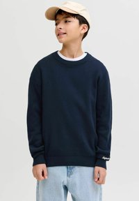 Garçon portant une casquette beige, un pull bleu marine, un sous-vêtement blanc et un jean bleu clair, regardant légèrement vers le haut et à droite.