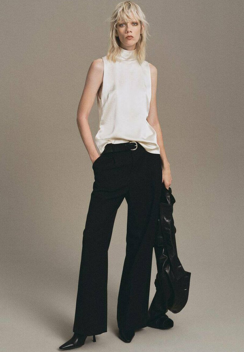 White sleeveless top with high neckline, paired with black wide-leg trousers. Black accessories include a handbag. Textured, sleek materials.