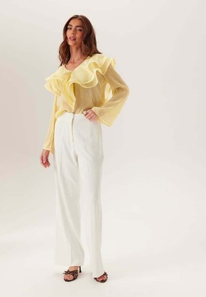 Kobieta ubrana w prześwitującą żółtą bluzkę z falbanami, białe spodnie z wysokim stanem i szerokimi nogawkami oraz sandały na wysokim obcasie w panterkę, stojąca na jasnym tle.