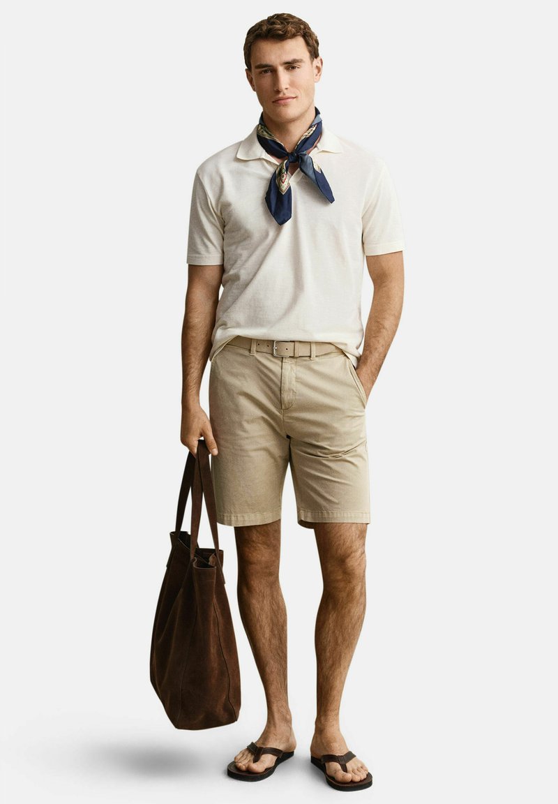Mann trägt weißes Polohemd, beige Shorts, navyfarbener Halstuch, braune Flip-Flops, hält große braune Tragetasche und steht mit einer Hand in der Tasche.