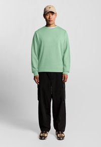 Mintgrön långärmad sweatshirt med rundad hals, tillsammans med svarta cargobyxor och mönstrade sneakers. Beigekaps med emblem på framsidan.