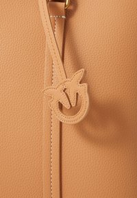 Tan krāsas ādas soma ar teksturētu virsmu, dekoratīvu šarmu ar ekscentrisku dizainu un baltām šuvēm gar jostu.