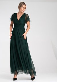 Langes grünes Kleid aus fließendem Stoff mit durchscheinenden, schwingenden Ärmeln und einem Wickeldesign. Der Saum hat ein durchscheinendes Overlay. Trägt dazu schwarze Pumps.