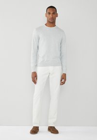 Suéter de punto gris, de mangas largas y cuello redondo; combinado con pantalones blancos y zapatos de ante marrón, de pie contra un fondo simple.