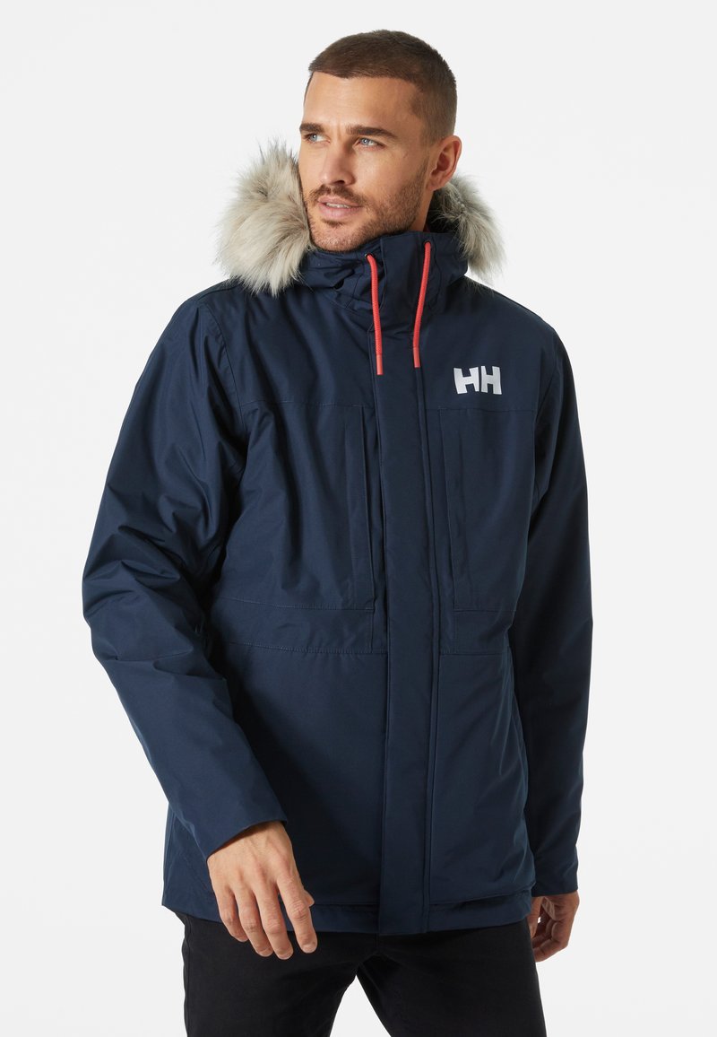 Chaqueta aislante de color azul marino con capucha forrada de piel sintética, cordones rojos, cierre de cremallera, dos bolsillos frontales y un logo en el pecho.