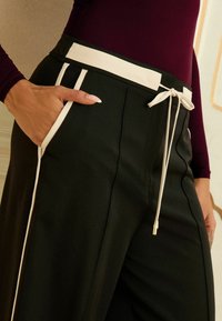 Zwarte broek met een witte tailleband en zijaccenten. Voorzien van een trekkoord en een zijzak. Gladde textuur en op maat gemaakte vorm.