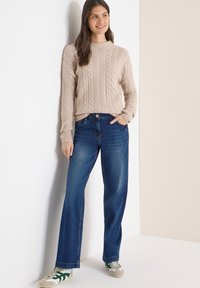 Femme appuyée contre un mur blanc, portant un pull beige en maille torsadée, un jean bleu et des baskets blanches à rayures vertes.