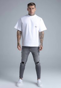 Fehér pamut rövid ujjú póló, szürke szakadt skinny farmerrel párosítva. A fehér tornacipő egészíti ki az öltözéket. Tetoválások láthatók a karokon.