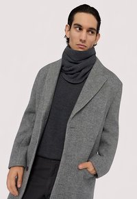 Šedý kabát se vzorem rybí kosti přes tmavě šedý svetr s tmavě šedým pleteným rolákem. Hladké textury a strukturovaný střih.